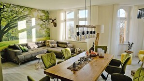 A cozy living room with a large window, a wooden dining table, and a green sofa.