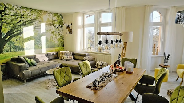 A cozy living room with a large window, a wooden dining table, and a green sofa. - "Tower Suite" - 130m² luxury suite: 3 bathrooms & bathtub (Bad Sachsa)