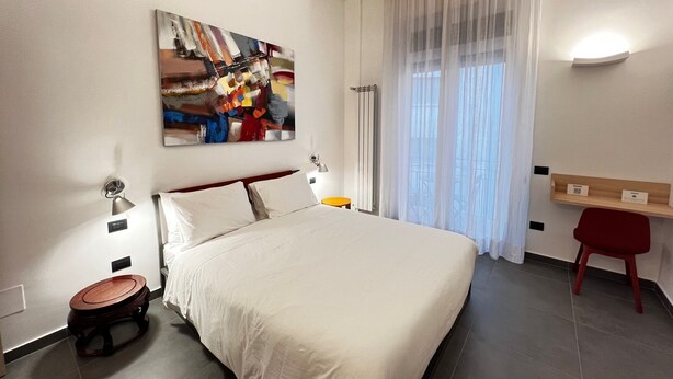 A modern hotel room with a large bed, a desk, a chair, a painting on the wall, and a window with curtains.