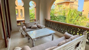 A balcony with a ceiling fan, a sofa, and a table. There are arched windows and a view of buildings and greenery. - Al Mahali Villas (Diani Beach)