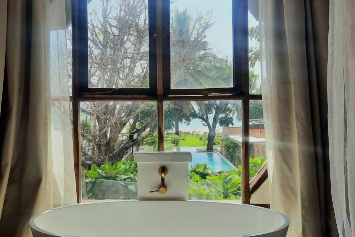 A bathroom with a bathtub, a window with curtains, and a view of a swimming pool and greenery.