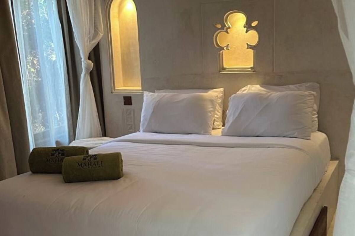 A neatly made bed with white linens and pillows, a wall-mounted light fixture, and a window with sheer curtains.