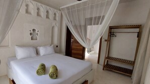 A neatly made bed with white linens and two rolled towels, a wooden bench, and a door leading to another room. - Al Mahali Villas (Diani Beach)
