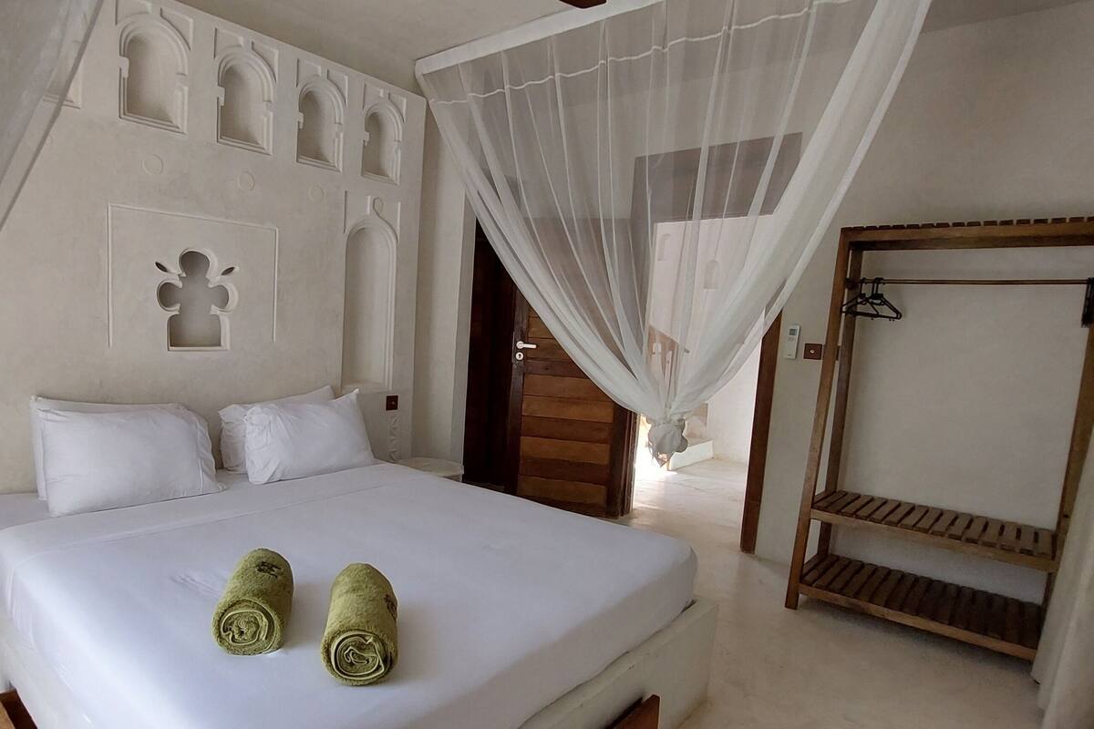 A neatly made bed with white linens and two rolled towels, a wooden bench, and a door leading to another room.