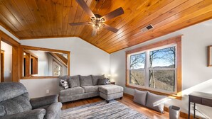 A modern living room with a sectional sofa, a ceiling fan, large windows, and a view of the outdoors.
