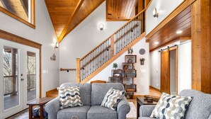 A spacious living room with a gray sofa set, wooden flooring, a staircase with a wooden railing, and a large wooden door.