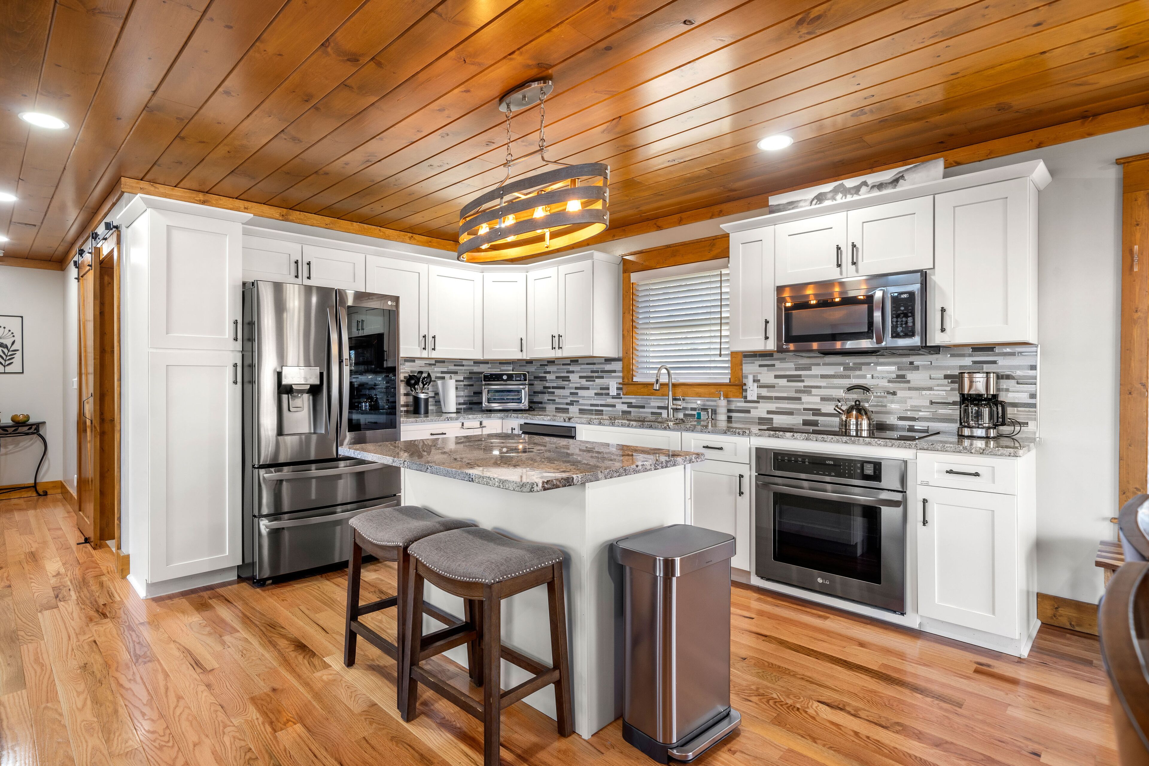 A modern kitchen with white cabinets, stainless steel appliances, a large island with stools, and a wooden ceiling.