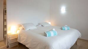 Un llit ben fet amb roba de llit blanca i dues tovelles, una taula de nit amb una làmpada, i un lluminari fixat a la paret.