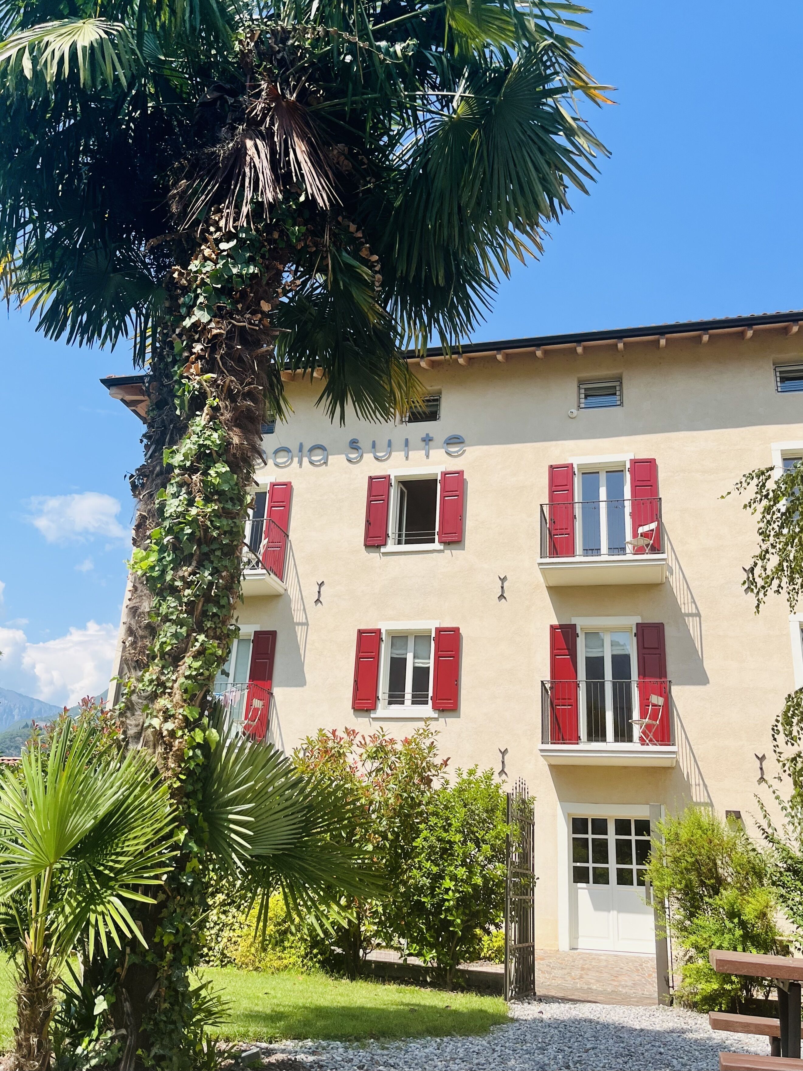 Egy épület, amelyen egy „villa apartman” feliratú tábla látható, piros zsaluzatos erkélyekkel, egy pálmafával és tiszta, kék éggel.
