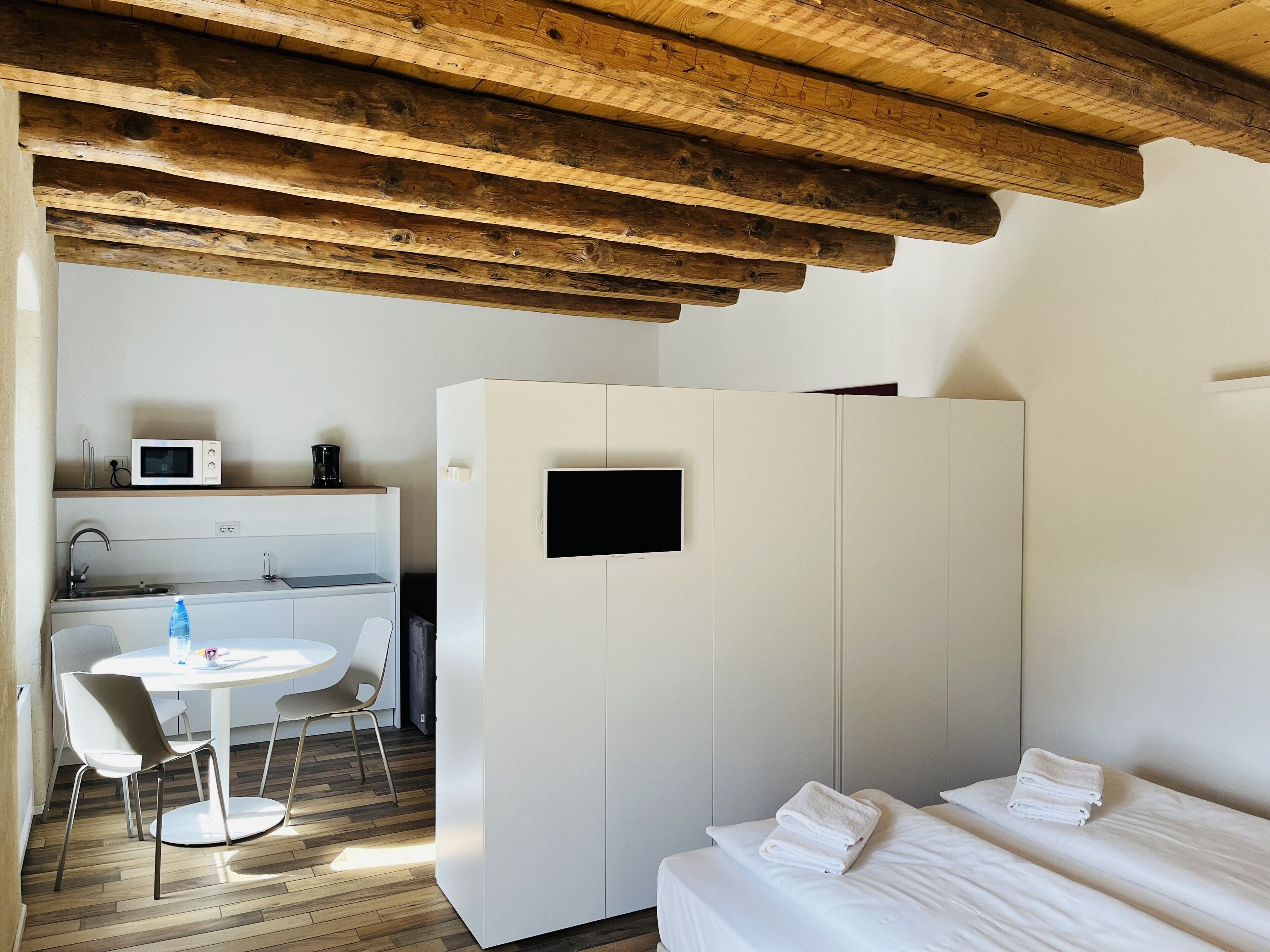 Ein modernes Zimmer mit Holzboden, einem weißen Kleiderschrank, einem Bett mit weißer Bettwäsche, einer kleinen Küchenzeile mit Mikrowelle und Kaffeemaschine sowie einem runden Tisch mit Stühlen.