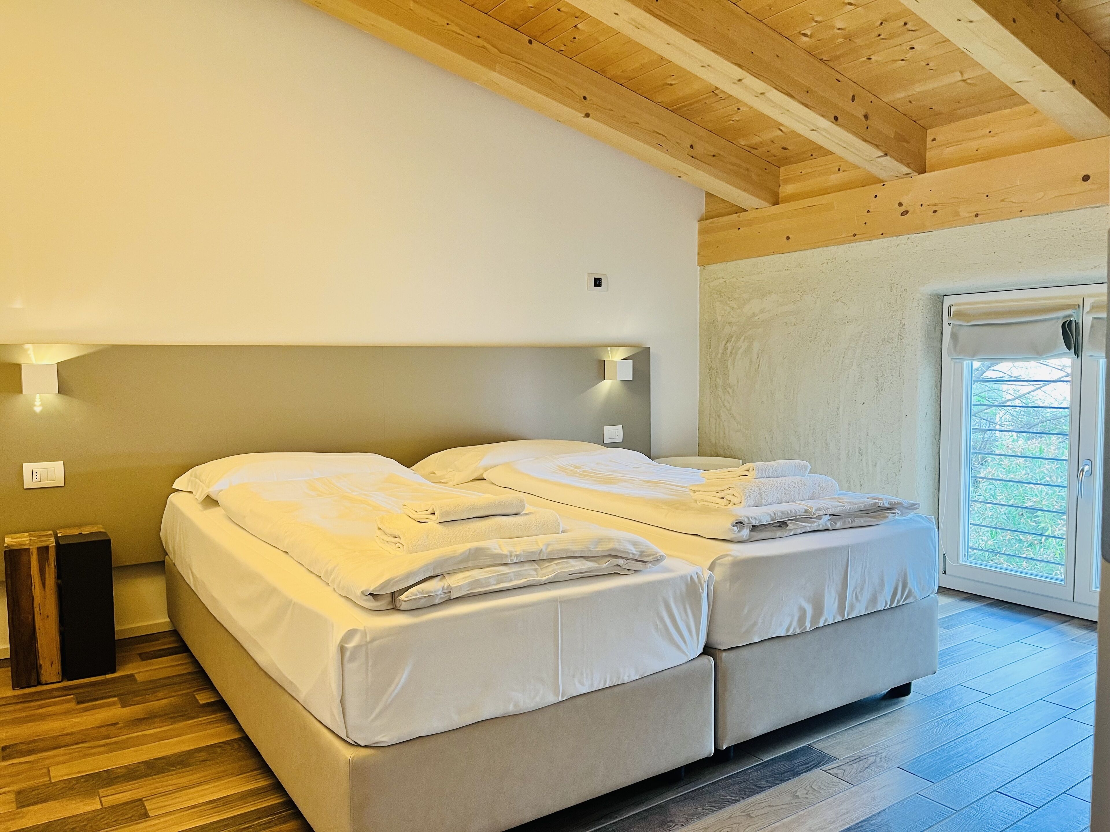 A double bed with white linens and towels, a wooden ceiling, a small bedside table, and a window with a view of greenery.