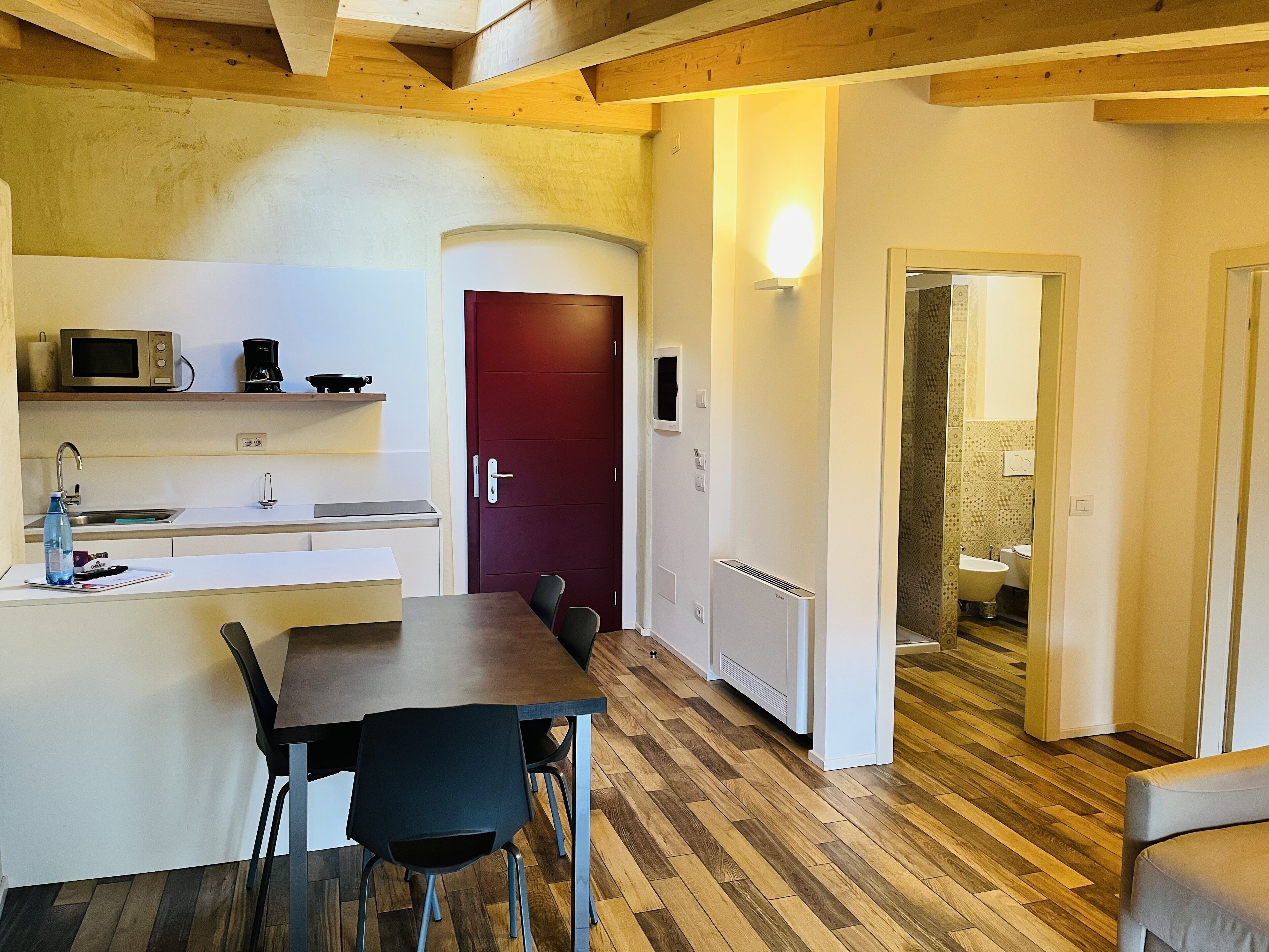 A modern kitchen with a dining area, wooden flooring, and a red door leading to a bathroom.