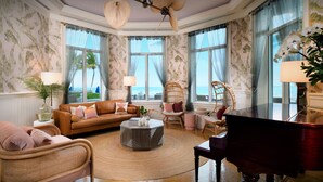 Amplia sala de estar con sofá, sillas, un piano y ventanas grandes con vista al mar.