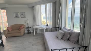 Een kamer met een bed, een bank, een bureau en een stoel. Er is een raam met gordijnen en uitzicht op de oceaan.