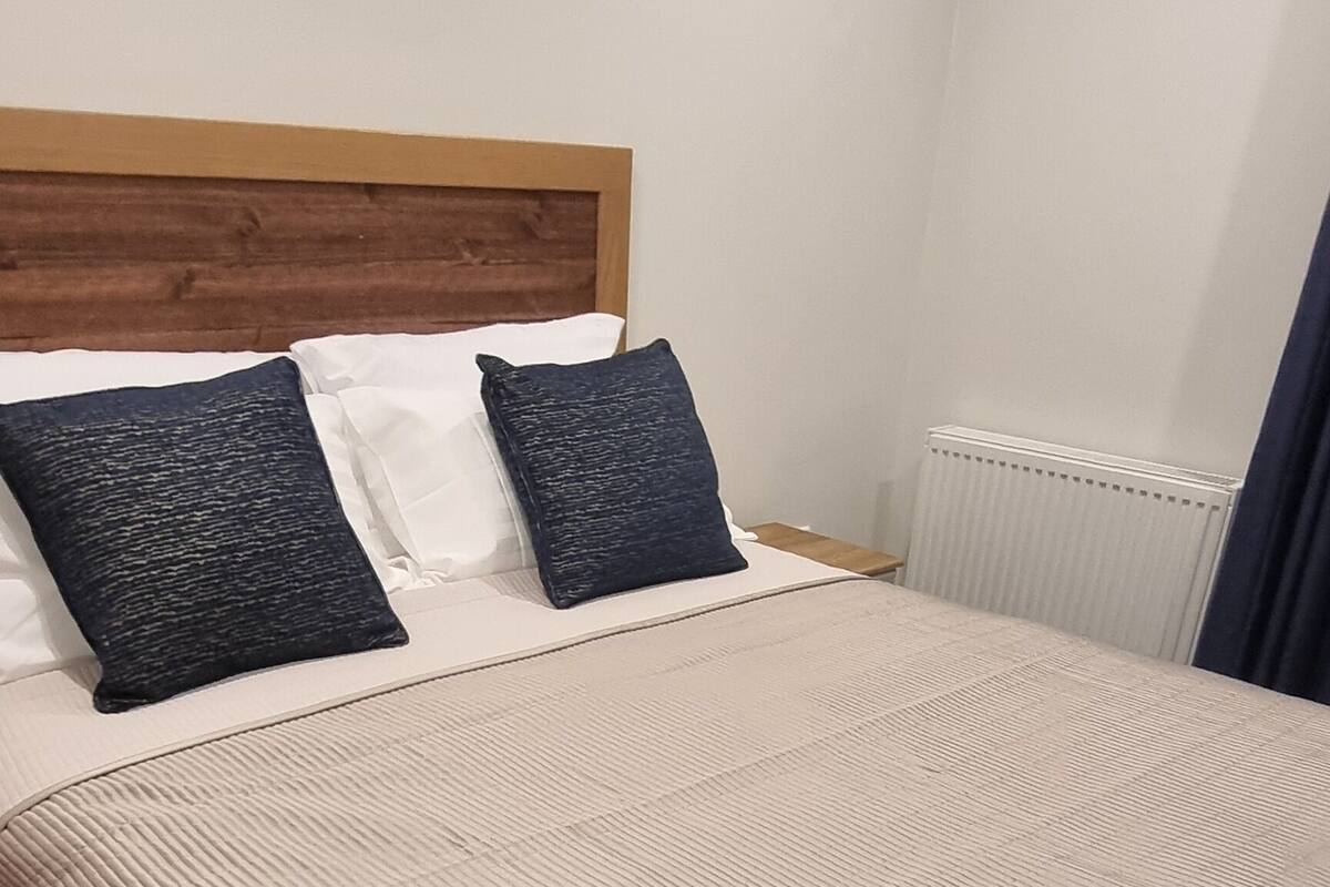 A neatly made bed with a beige quilt and two dark pillows, a wooden headboard, and a radiator in the corner.