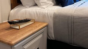 Una cama bien tendida con almohadas blancas y azules, un control remoto sobre una mesita de noche de madera y un cajón de mesita de noche blanco.