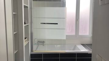 A modern bathroom with a glass shower enclosure, a white bathtub, and a window with frosted glass.