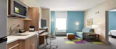 A compact hotel room with a kitchenette, a sofa, a desk, and a television.