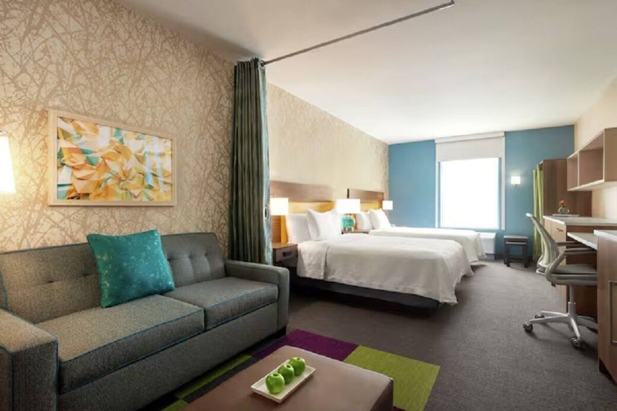 A hotel room with a sofa, two beds, a desk, and a painting on the wall.