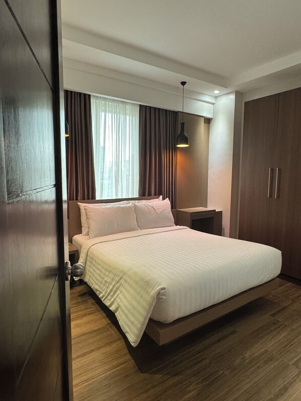 A modern bedroom with a large bed, wooden flooring, a nightstand, a wardrobe, and a window with curtains.