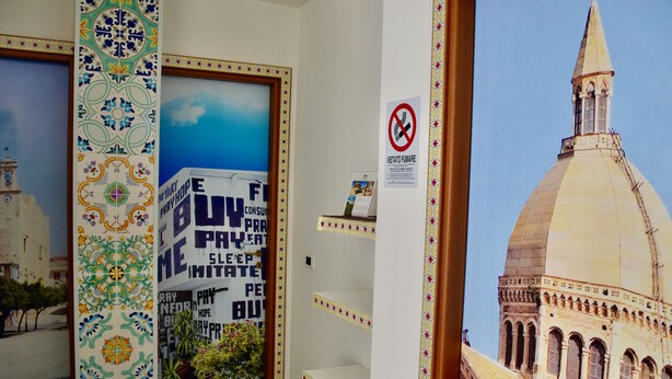 Un couloir orné d’une colonne à motifs, une porte avec une affiche, un panneau «interdiction de fumer» et une vue sur un bâtiment coiffé d’un dôme.