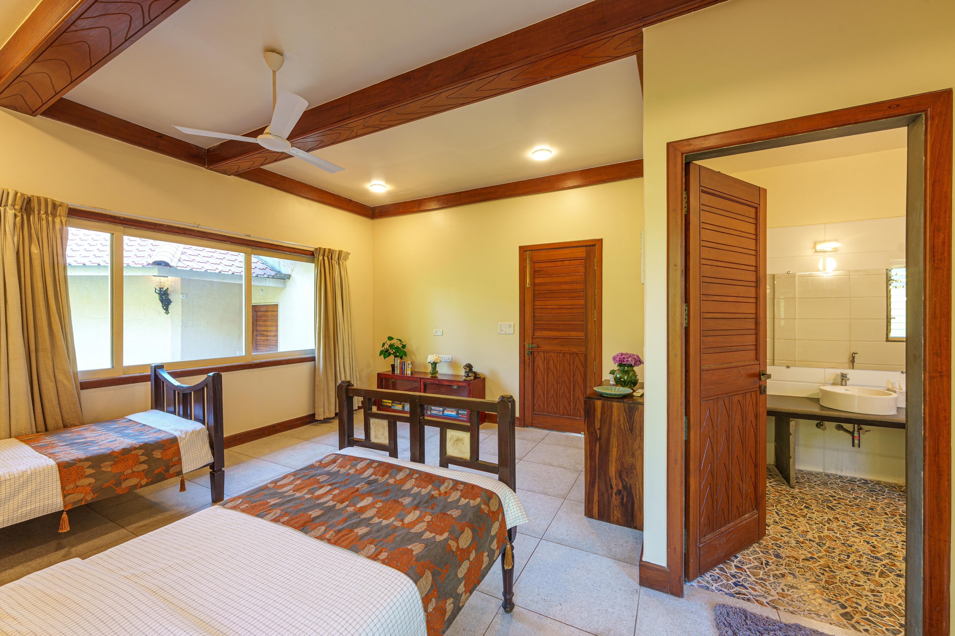 A hotel room with two beds, a ceiling fan, a wooden door, a bathroom with a sink and mirror, and a window with curtains.