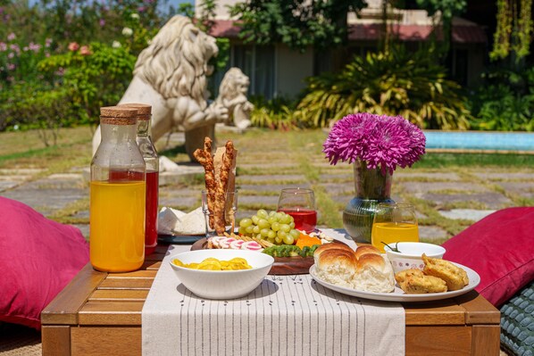 A picnic setup with food and drinks on a wooden table outdoors, surrounded by greenery and a statue.