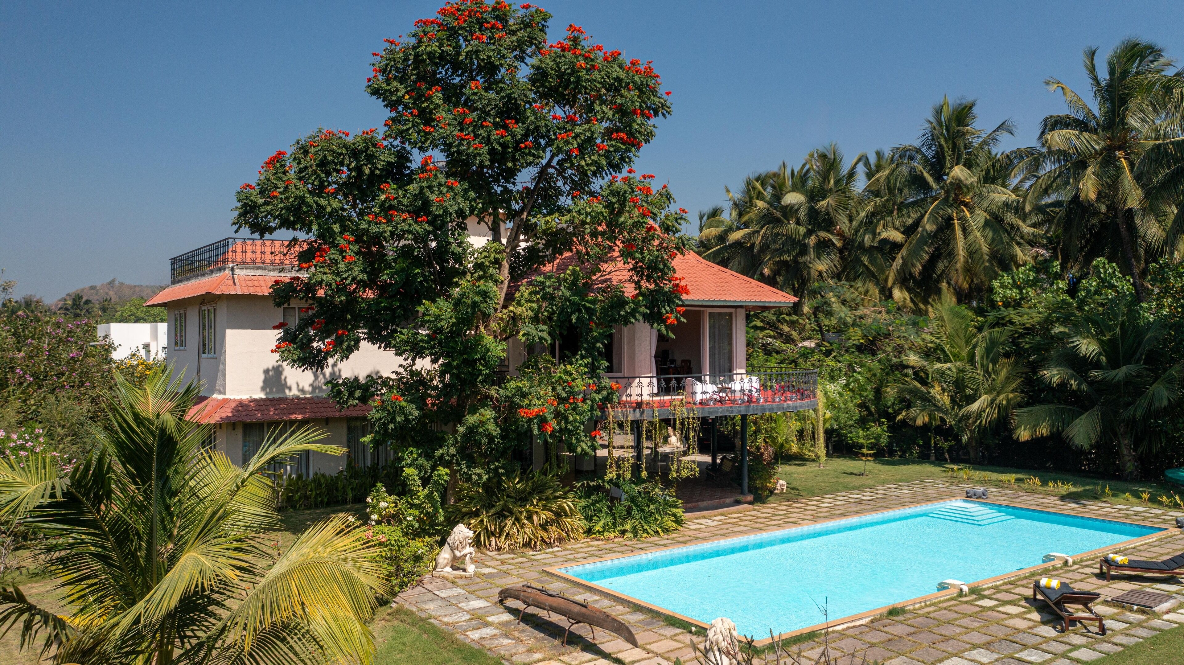 A two-story house with a red roof, a swimming pool, and a garden with palm trees and flowering plants.