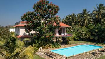 A two-story house with a red roof, a swimming pool, and a garden with palm trees and flowering plants.