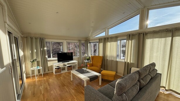Een ruime woonkamer met een groot raam, houten vloeren, een grijze bank, een geel fauteuil en een televisie op een witte standaard.
