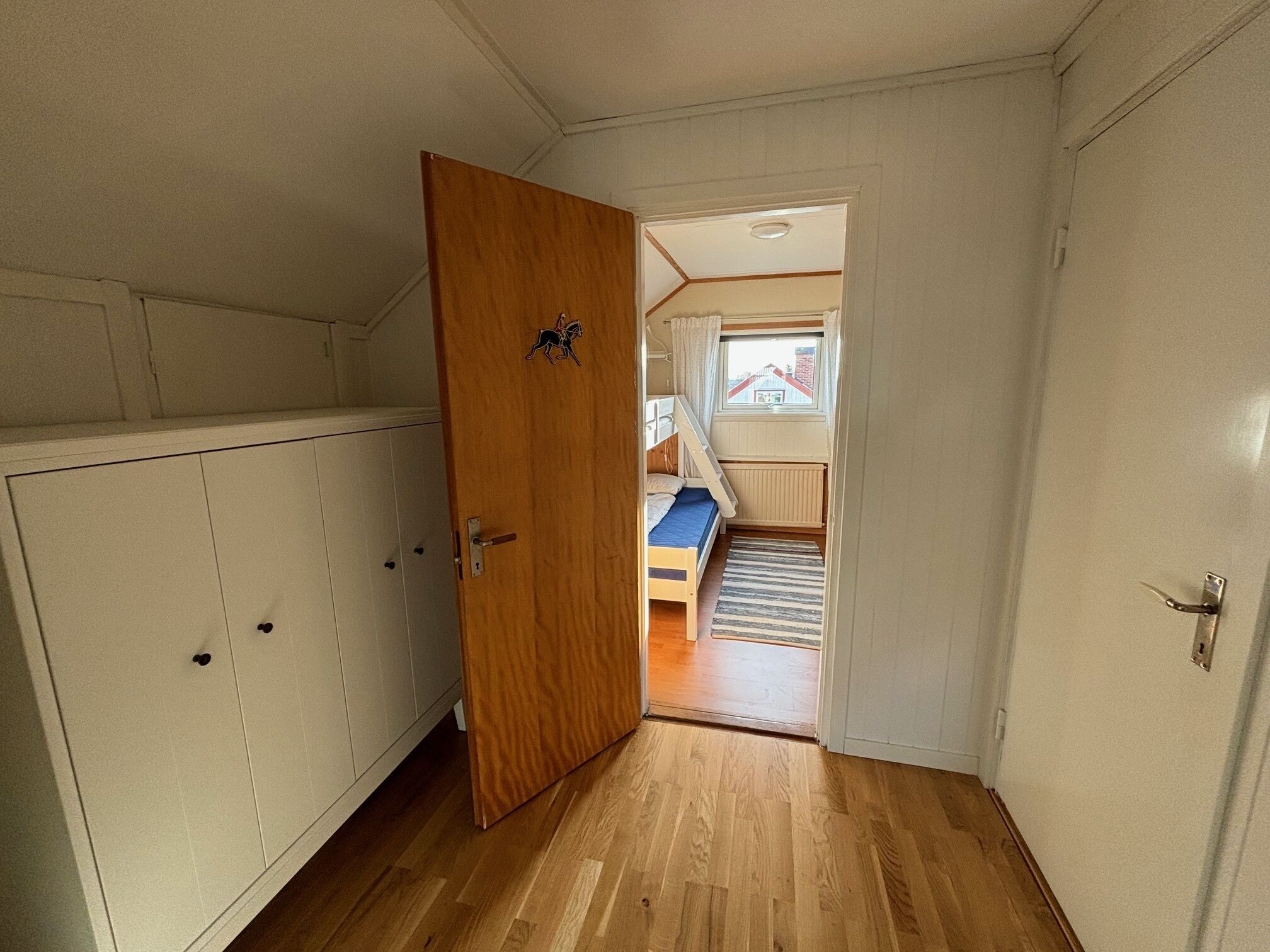 Un couloir étroit avec une porte en bois qui donne sur une pièce où se trouve un lit superposé et une fenêtre.