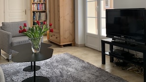 Un salon avec un canapé gris, une table basse ornée d’un vase de tulipes rouges, un meuble en bois, un téléviseur posé sur un meuble, et une grande fenêtre donnant sur les bâtiments environnants.