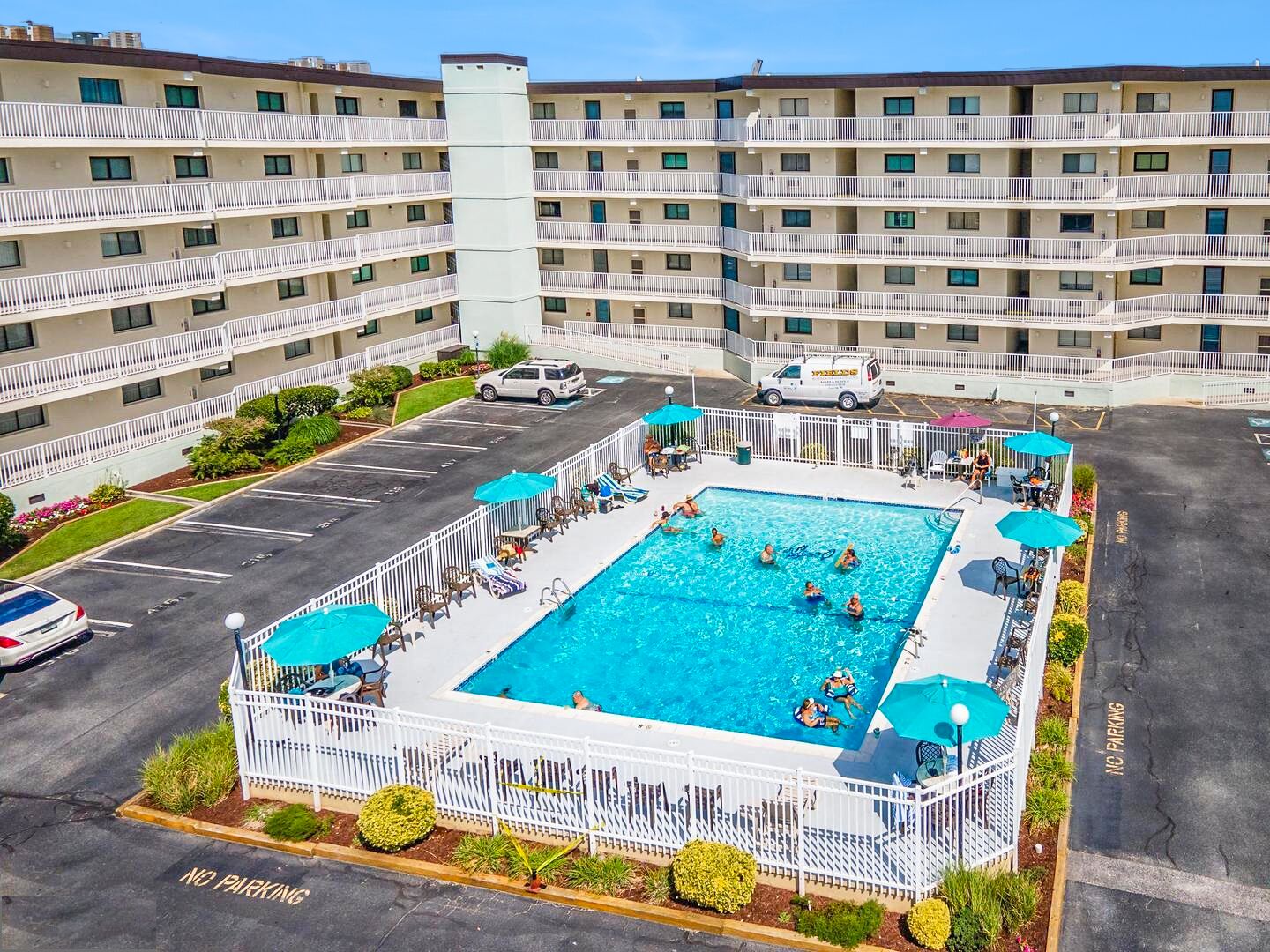 A hotel with a central swimming pool surrounded by a white fence, multiple balconies, and a parking area.
