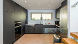 A modern kitchen with dark cabinetry, a stainless steel refrigerator, a dishwasher, and a window above the sink. - SeaWhisper Villa - Four Bedroom Villa, Sleeps 8 (Paxi)