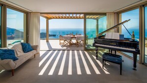 A modern living room with a piano, a sofa, and a dining area with a view of the sea. - SeaWhisper Villa - Four Bedroom Villa, Sleeps 8 (Paxi)