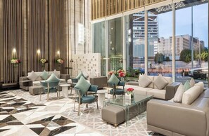 Un hall d’entrée moderne avec un sol à motifs géométriques, un espace détente doté de canapés et de fauteuils, et de grandes fenêtres offrant une vue sur le paysage urbain.