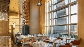 A modern restaurant with a high ceiling, exposed beams, and large windows offering a city view.
