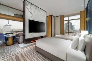 A modern hotel room with a large bed, a flat-screen TV, a fireplace, a city view, and a balcony with a table and chairs.