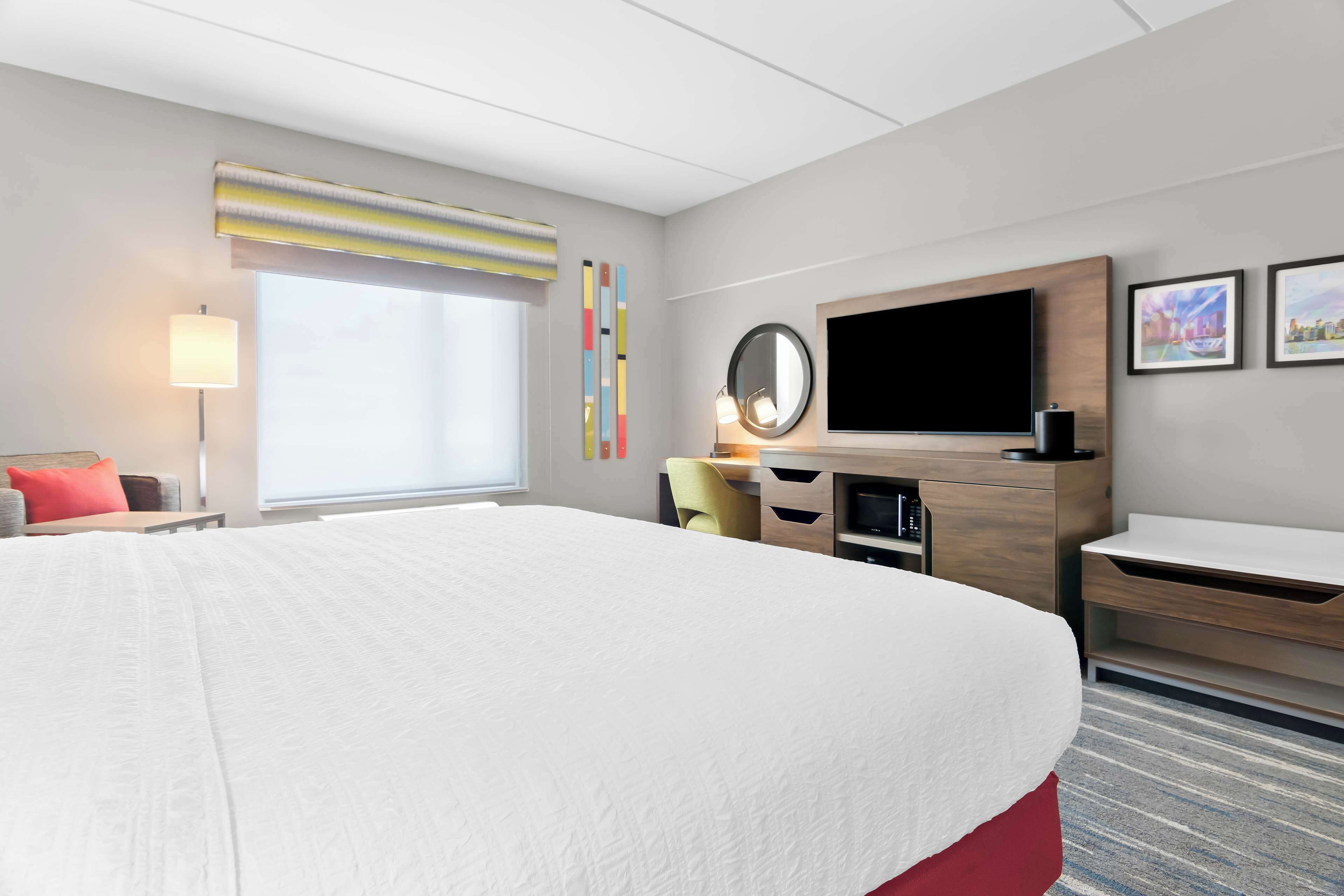 Ein Hotelzimmer mit einem großen Bett, einem Schreibtisch mit Computer, einem Flachbildfernseher und einem Fenster mit Jalousien.