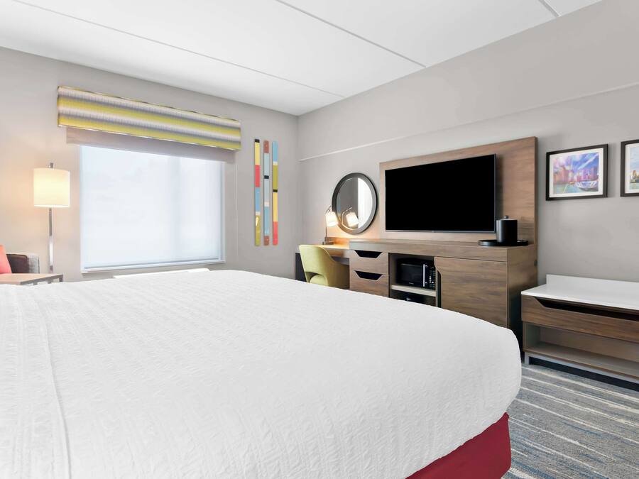 A hotel room with a large bed, a desk with a computer, a flat-screen TV, and a window with blinds.