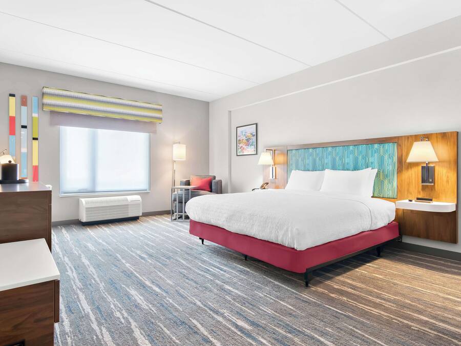 Habitación de hotel con una cama grande, un escritorio, una silla, una ventana con persianas, un televisor montado en la pared y una mesita de noche con lámpara.