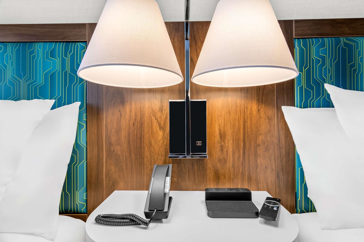 Ein Hotelzimmer mit einem hölzernen Kopfteil, zwei großen Lampen, einem Telefon, einer Ladestation und einem kleinen Tisch mit einer Fernbedienung.