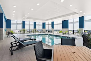 A modern indoor pool area with lounge chairs, a wooden table, and large windows.