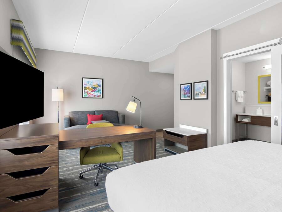 Una habitación de hotel con cama, escritorio, silla, televisor, lámpara y obras de arte en las paredes.