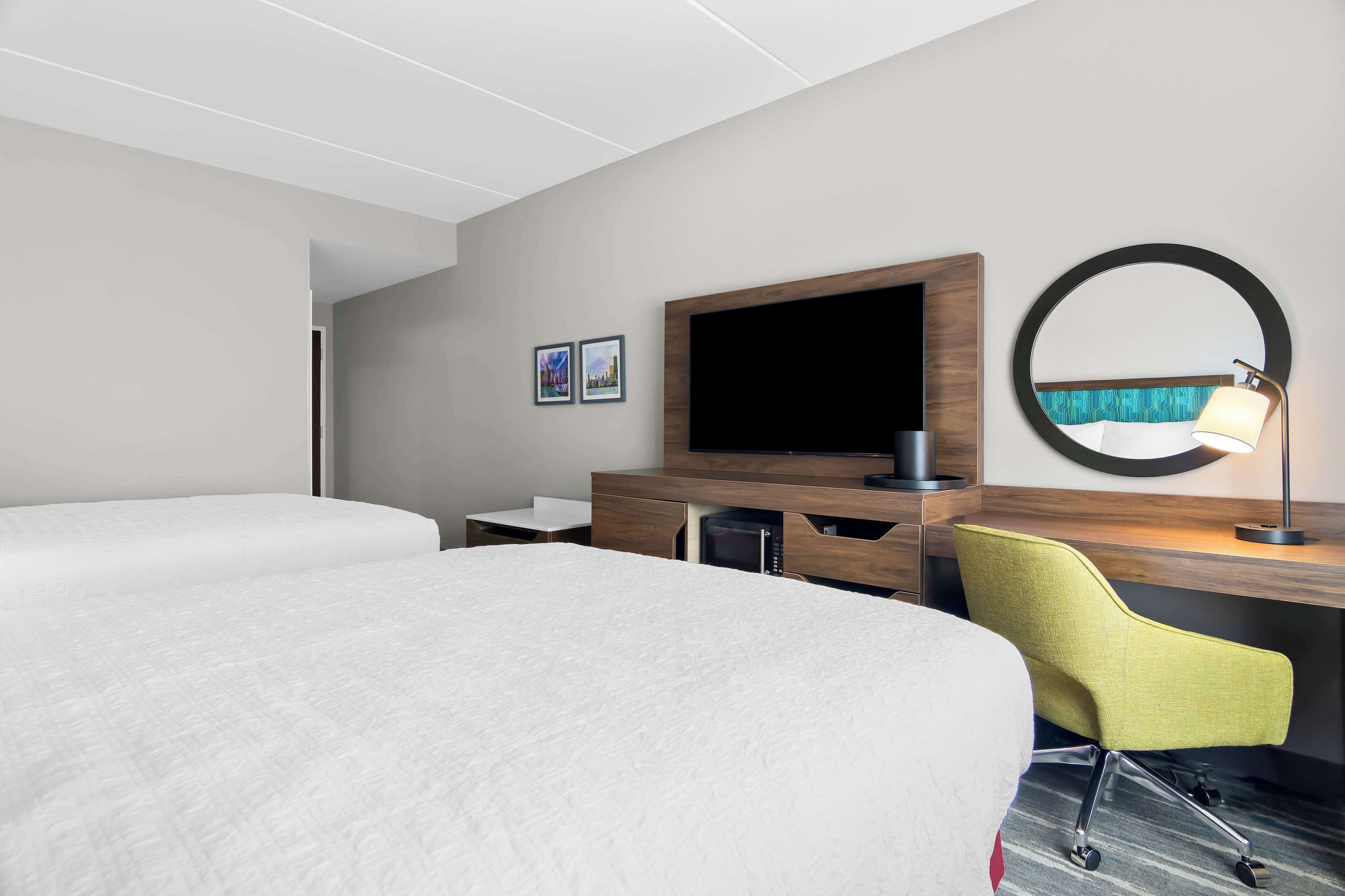 A hotel room with two beds, a desk with a chair, a television, a mirror, and a lamp.
