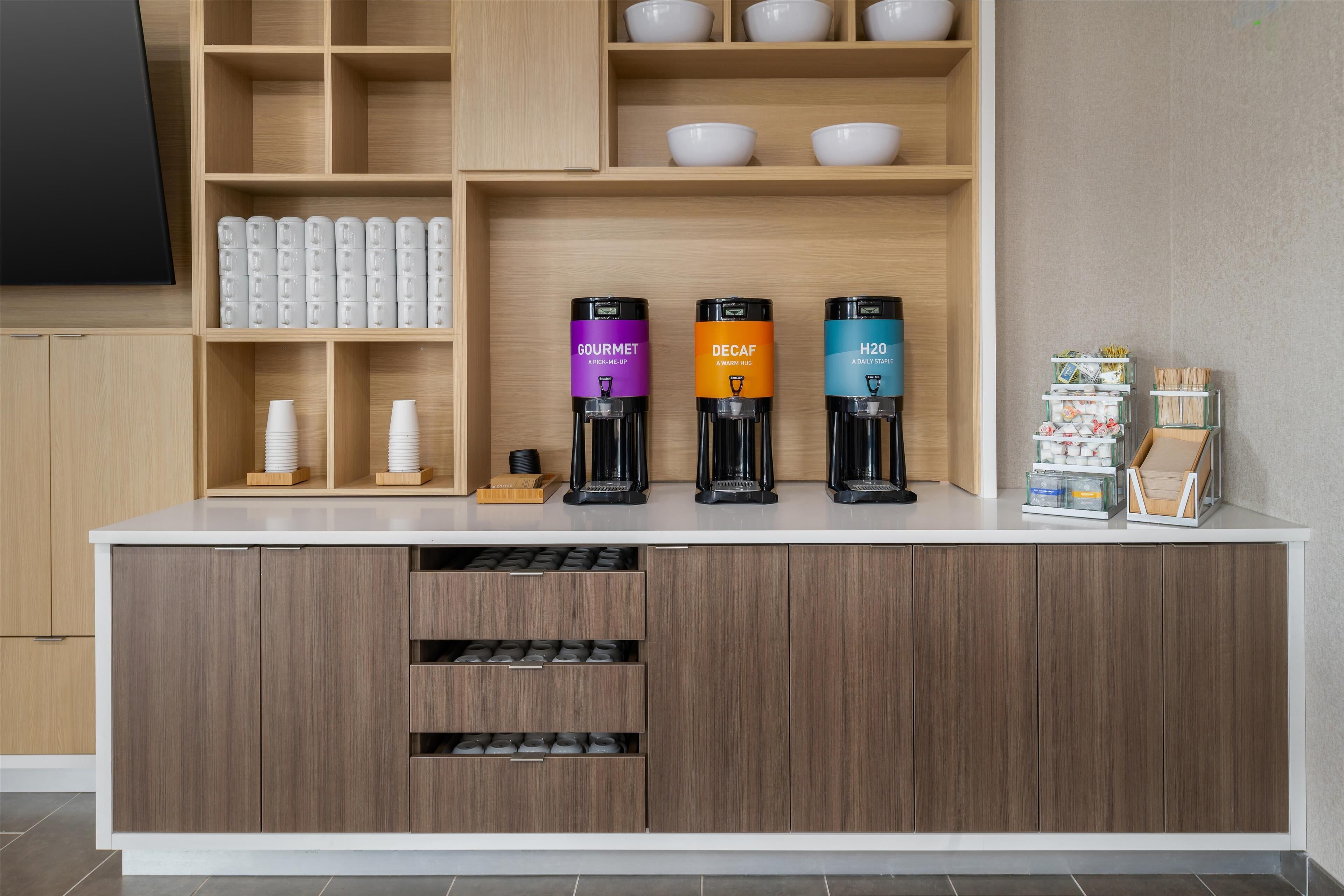 A modern kitchen with a coffee station featuring three coffee dispensers labeled 'GOURMET,' 'DECAF,' and 'H2O.' There are white cups and bowls on shelves, and a small display of snacks and condiments.