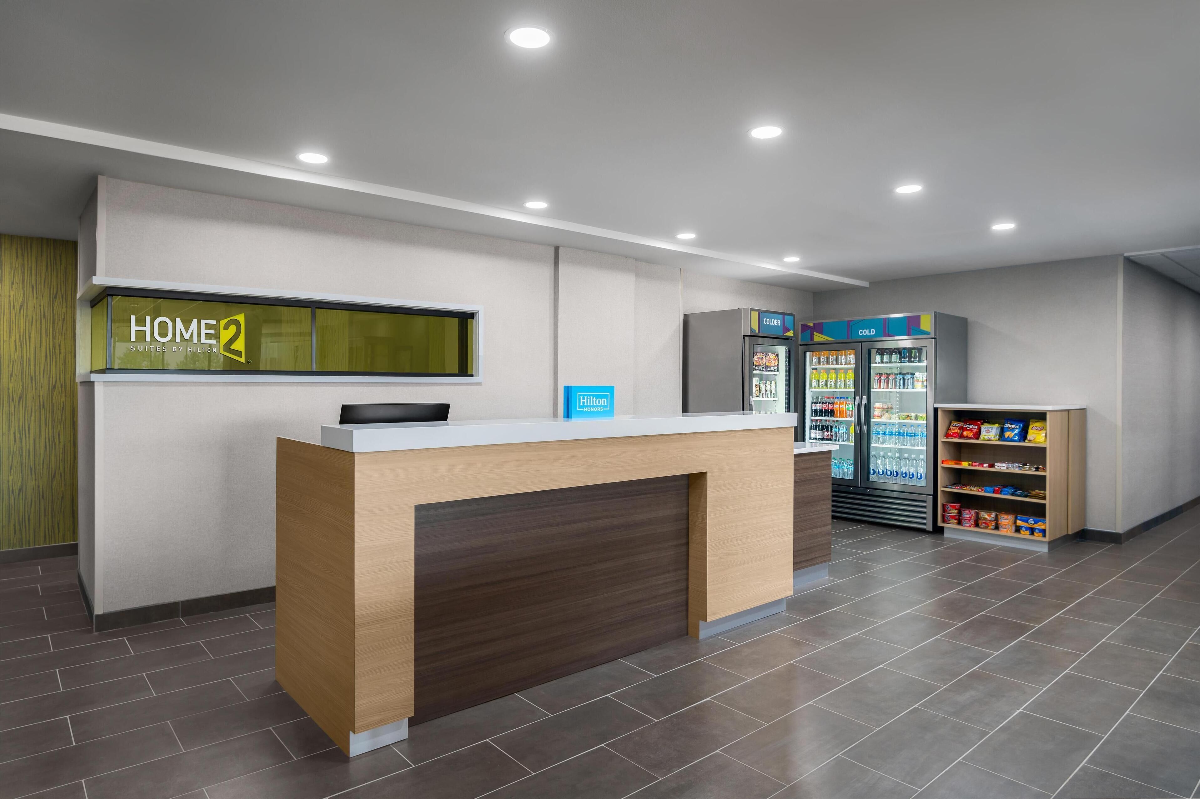 A hotel reception area with a reception desk, a 'Home 2 Suites by Hilton' sign, a minibar, and a refrigerator with beverages.