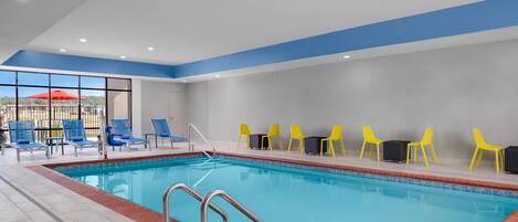 An indoor swimming pool with blue and yellow lounge chairs, a tiled floor, and a view through large windows.