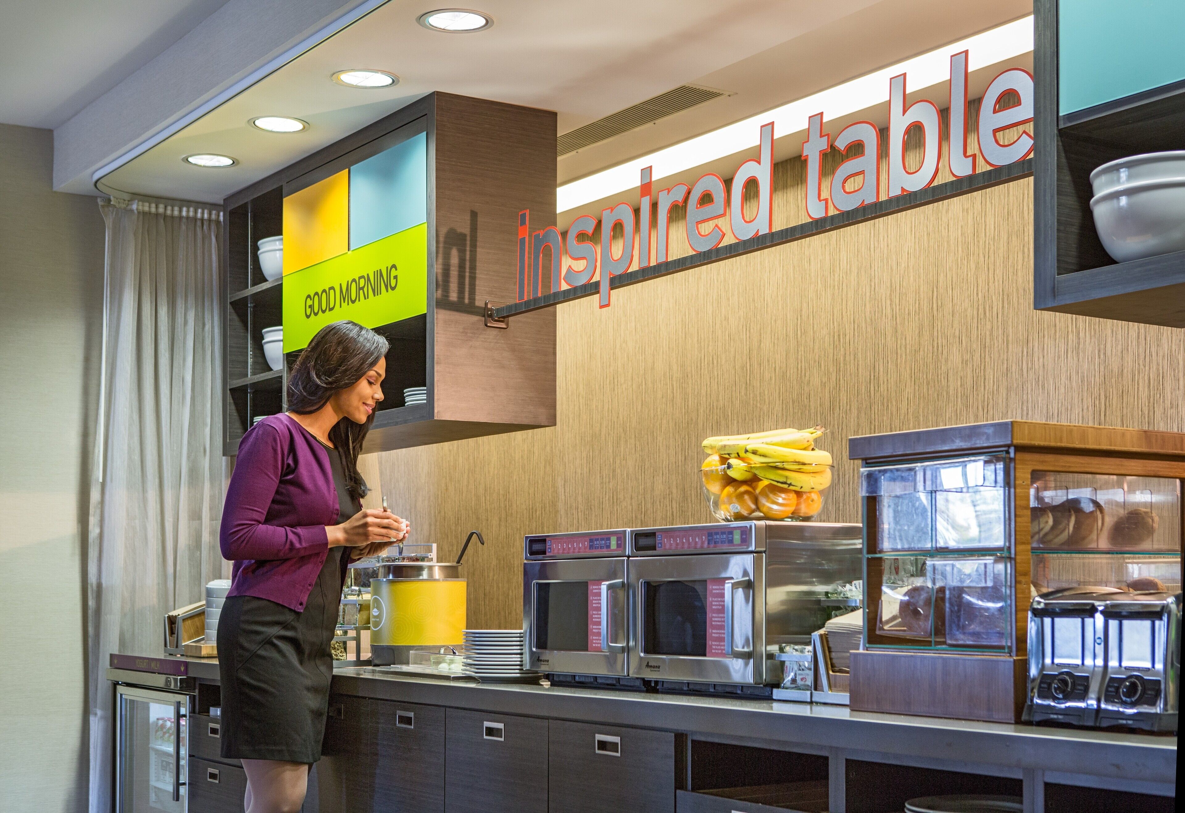 A breakfast bar with a sign reading "inspired table," a microwave, toaster, and a bowl of fruit.