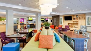 Een moderne hotellobby met een centraal zitgedeelte, een keuken op de achtergrond en een televisie aan de muur.